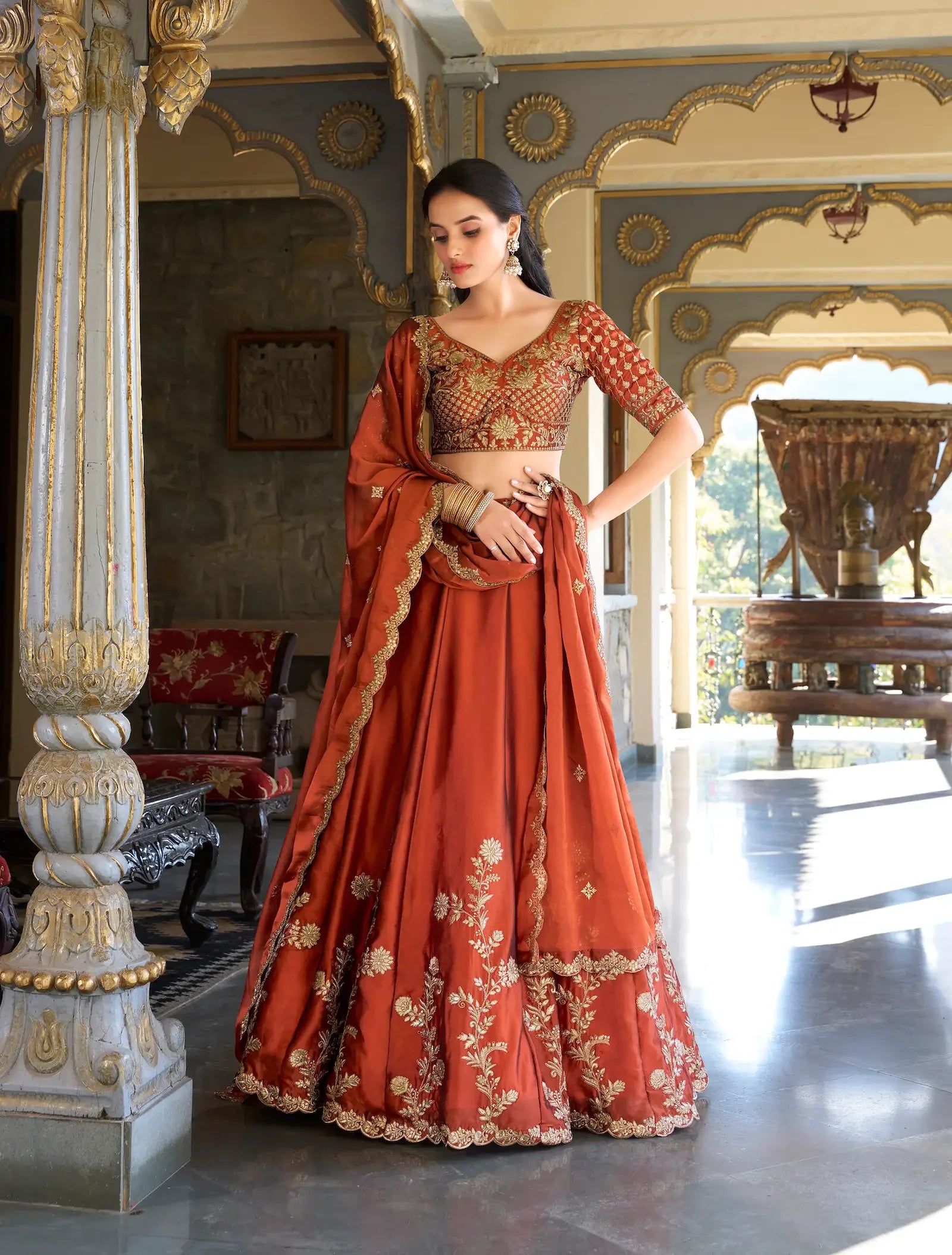 Woman in an embroidered burnt orange lehenga with gold details, standing in a palace hallway