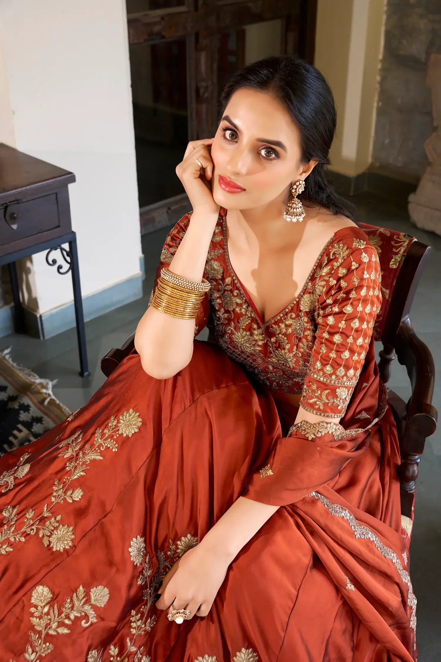 Woman in a rust-red embroidered lehenga with gold jewelry seated indoors