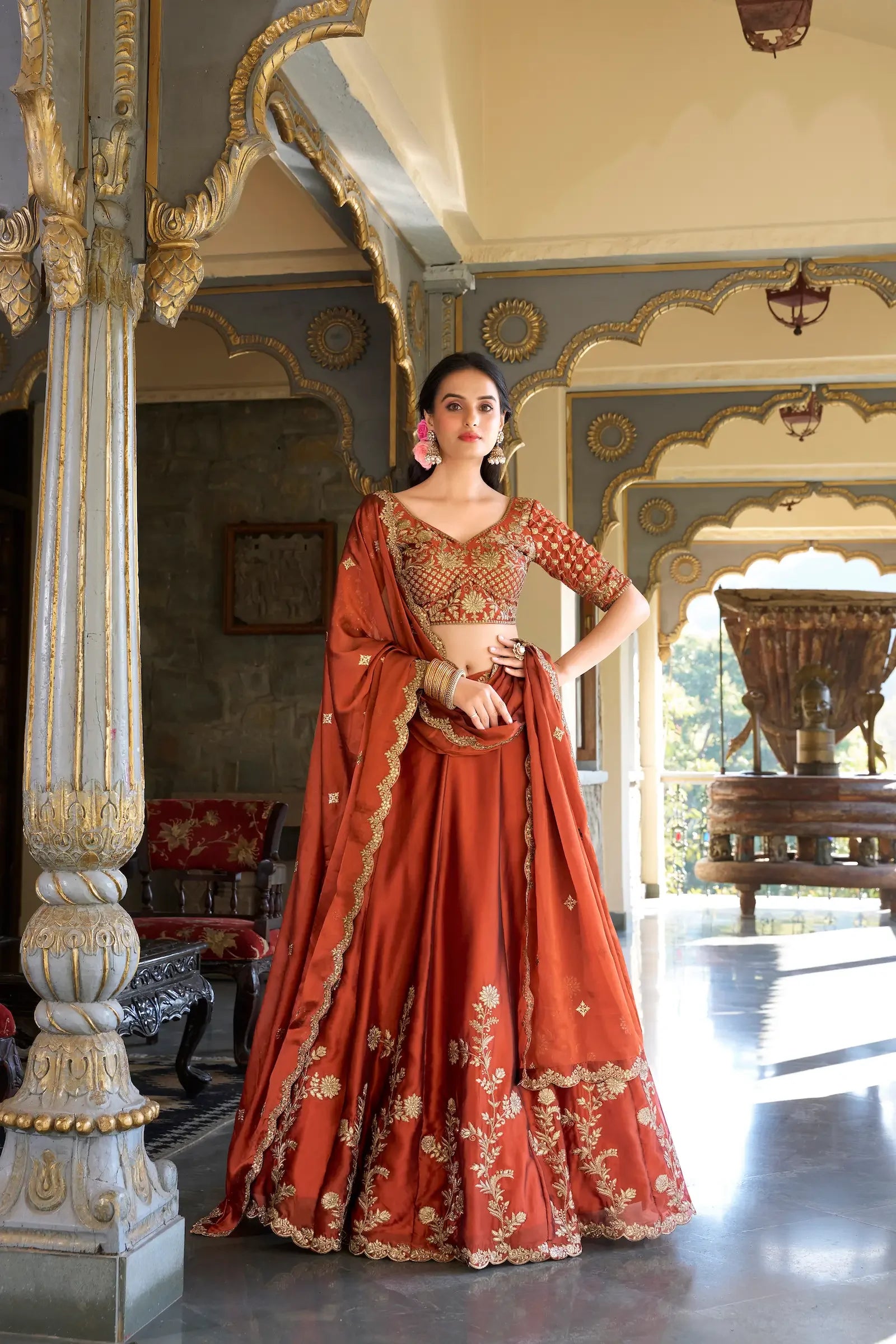 Woman in ornate rust orange lehenga with gold embroidery standing in traditional interior