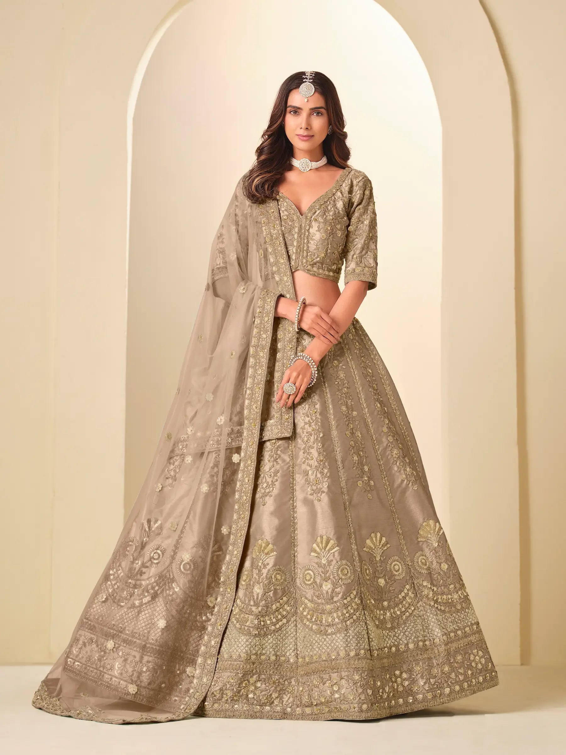 Woman in beige embroidered lehenga choli with dupatta, standing in arched setting