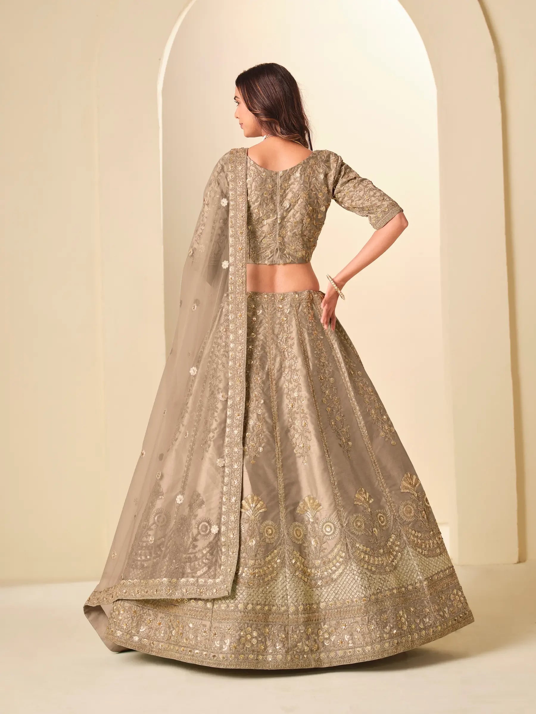 Woman in gold embroidered lehenga with dupatta, standing against arched neutral background