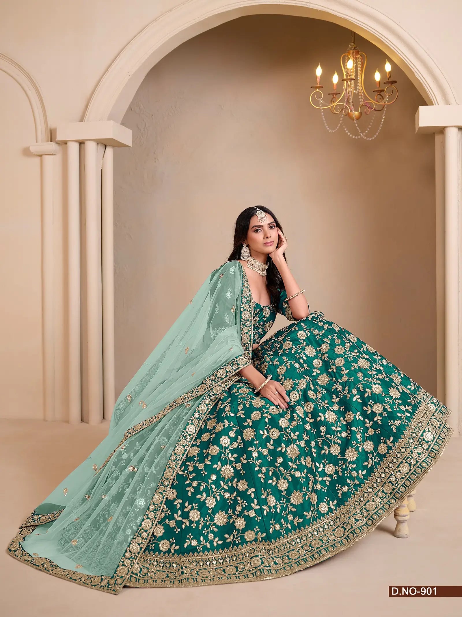 Woman in an embroidered green lehenga with gold floral patterns and pastel dupatta, sitting indoors under a chandelier.