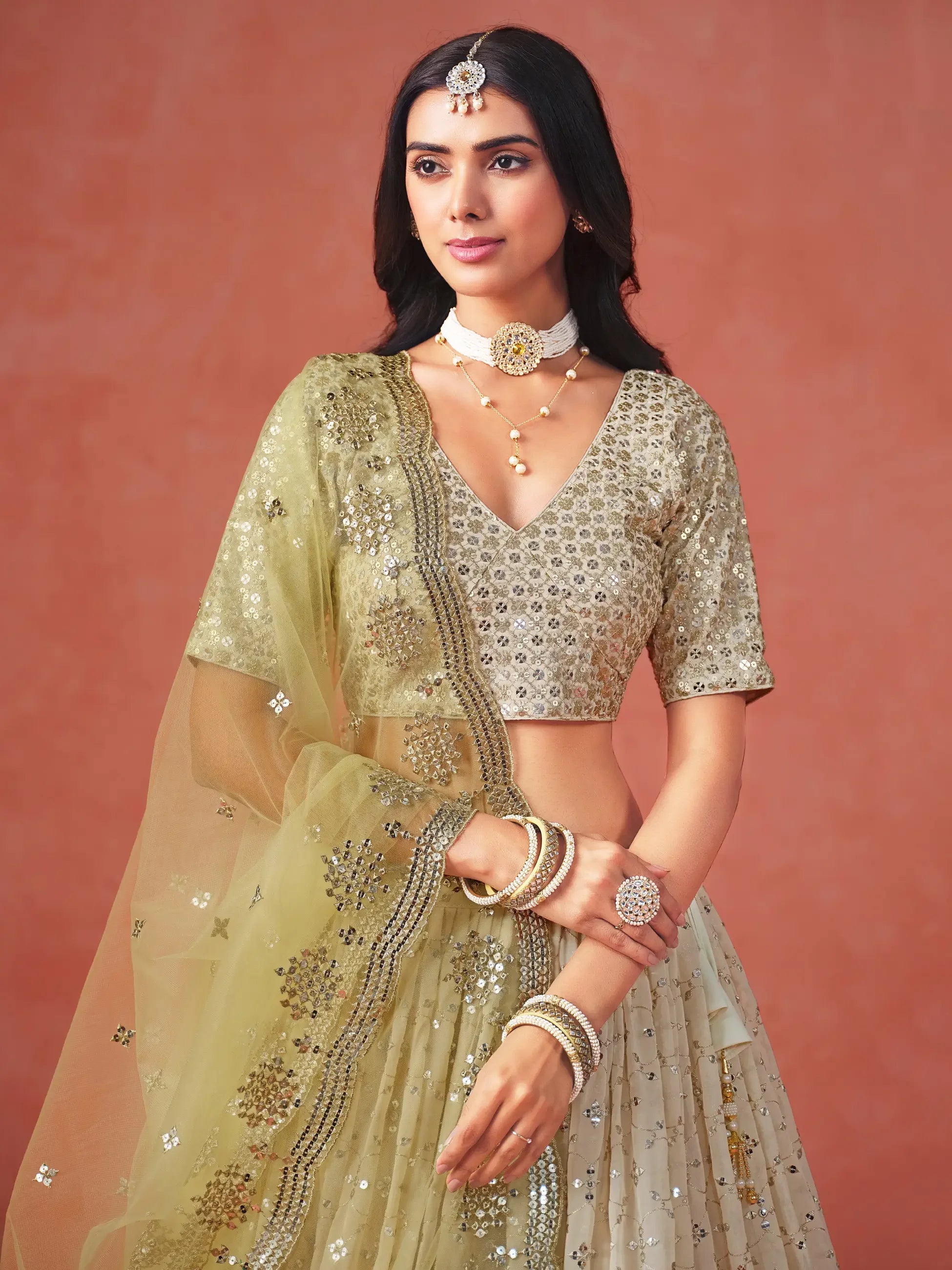 Woman in embellished lehenga choli with dupatta, traditional jewelry, and pastel backdrop