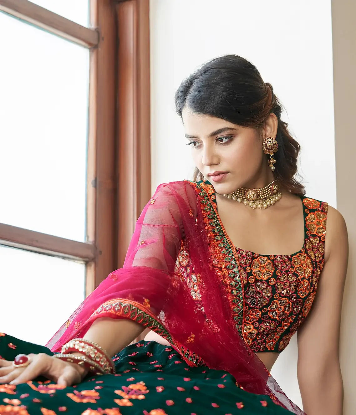 Woman in traditional Indian attire with embroidered blouse and pink net dupatta, sitting by window