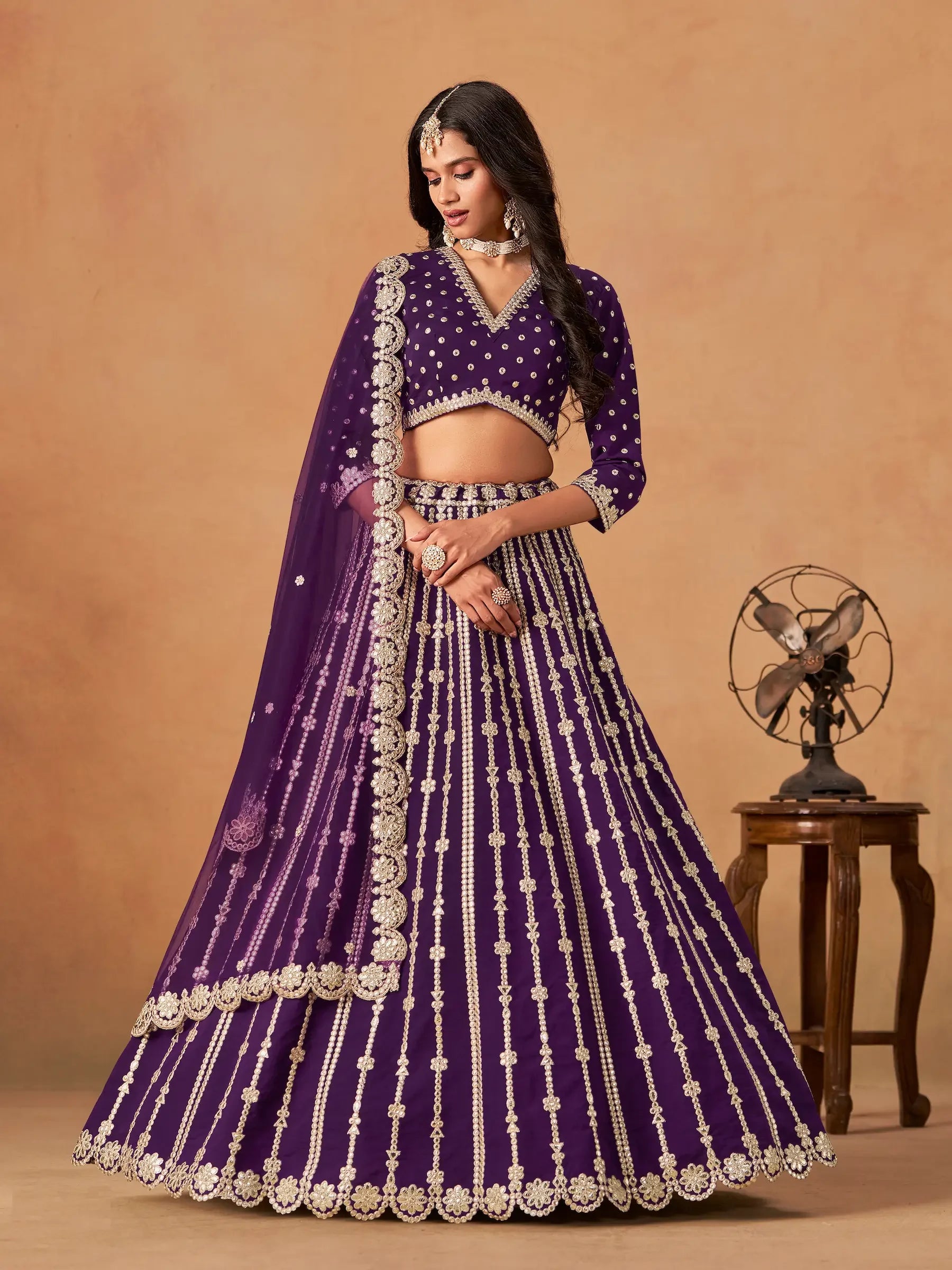 Woman wearing a purple embroidered lehenga with gold accents, traditional jewelry, and dupatta.