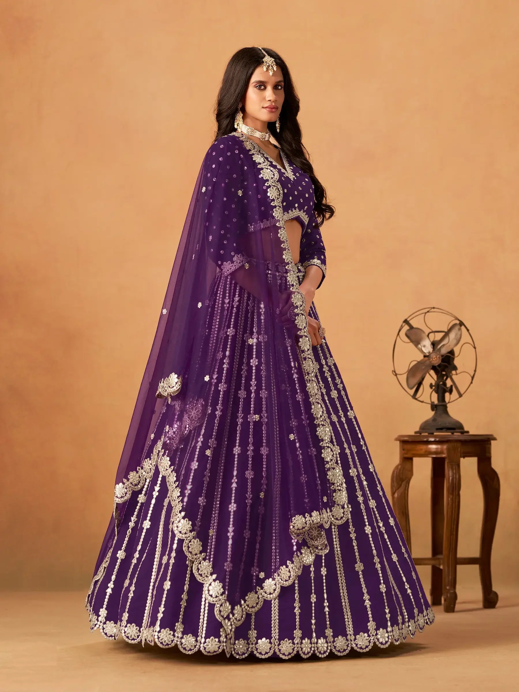 Woman wearing a purple embroidered lehenga with dupatta, standing indoors beside a table fan.