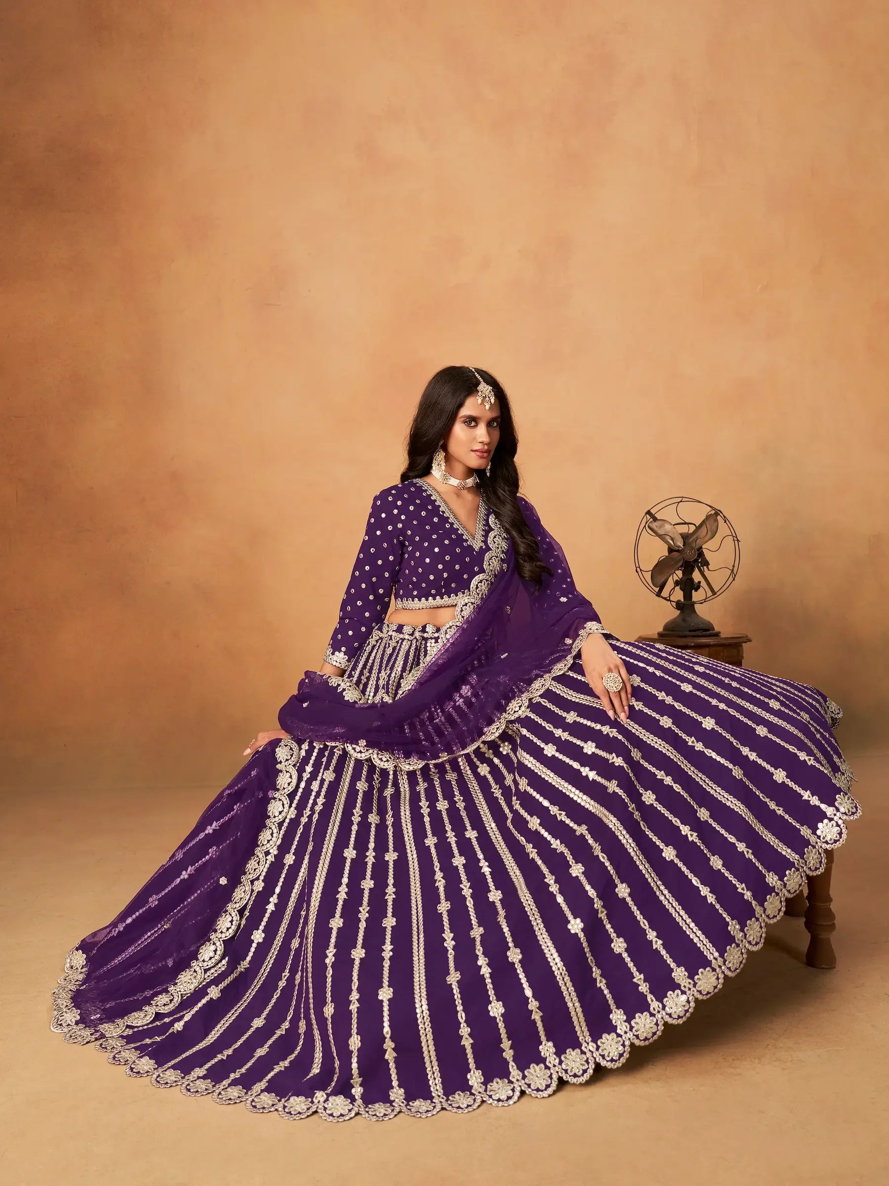 Woman in purple embroidered lehenga set with silver jewelry, sitting in studio setting