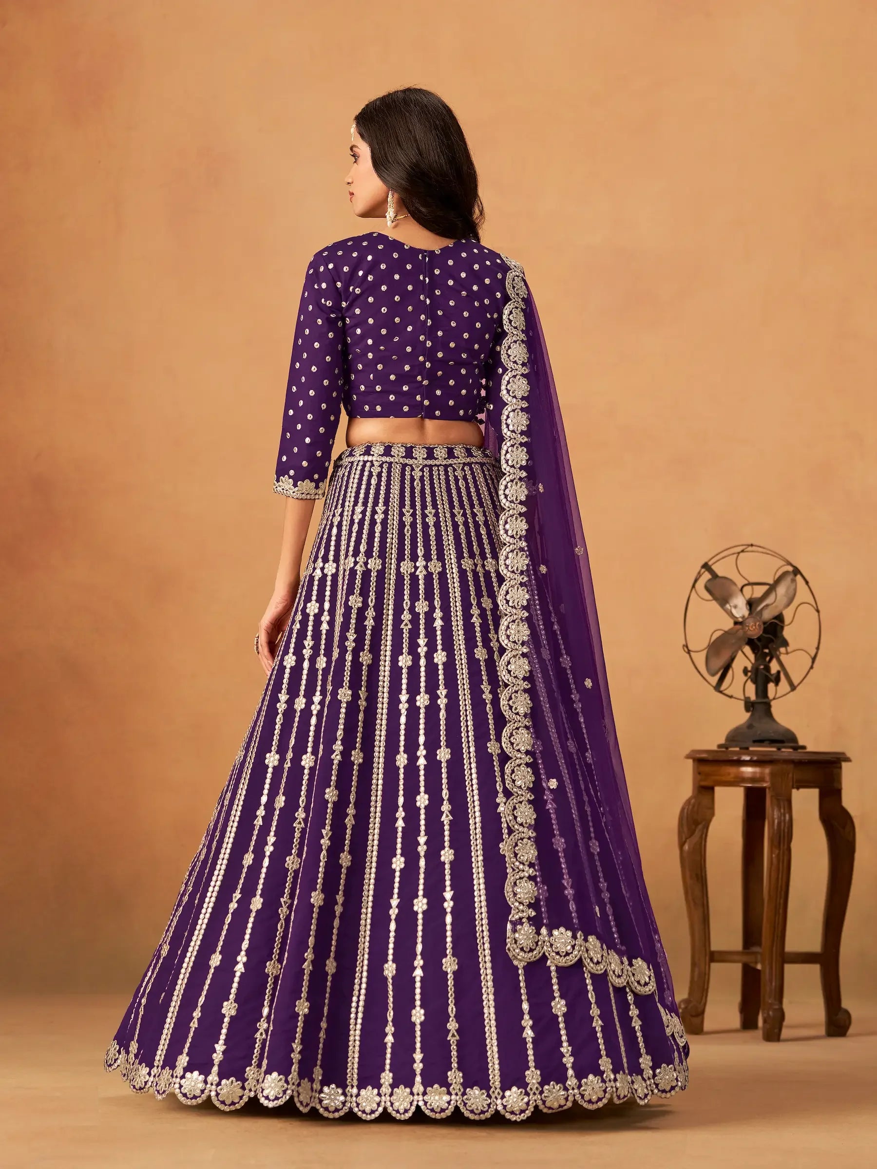 Woman wearing a purple embroidered lehenga set, shown from the back, with dupatta and silver details.
