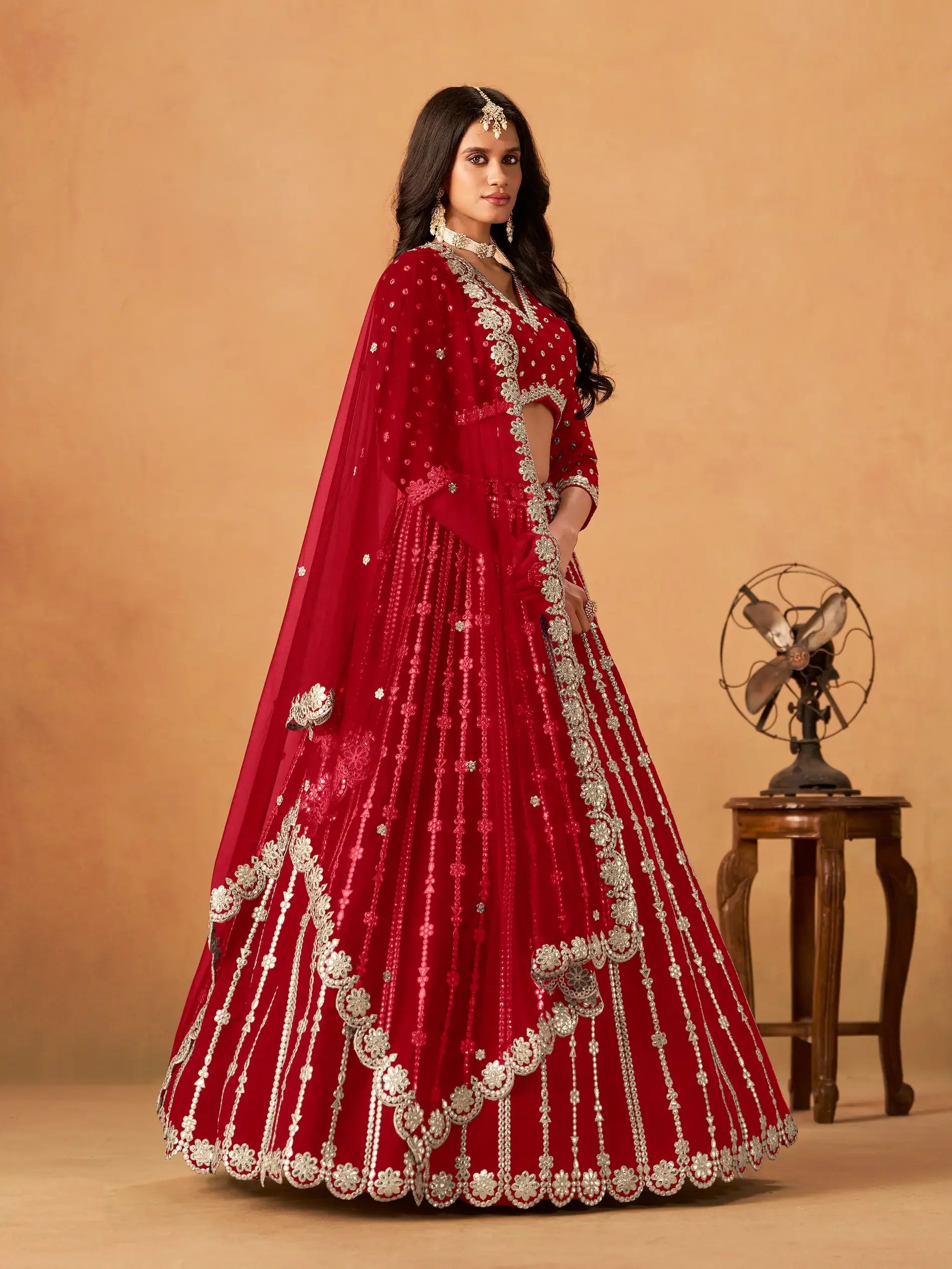 Woman wearing red embroidered lehenga with silver details, posing indoors next to a vintage fan