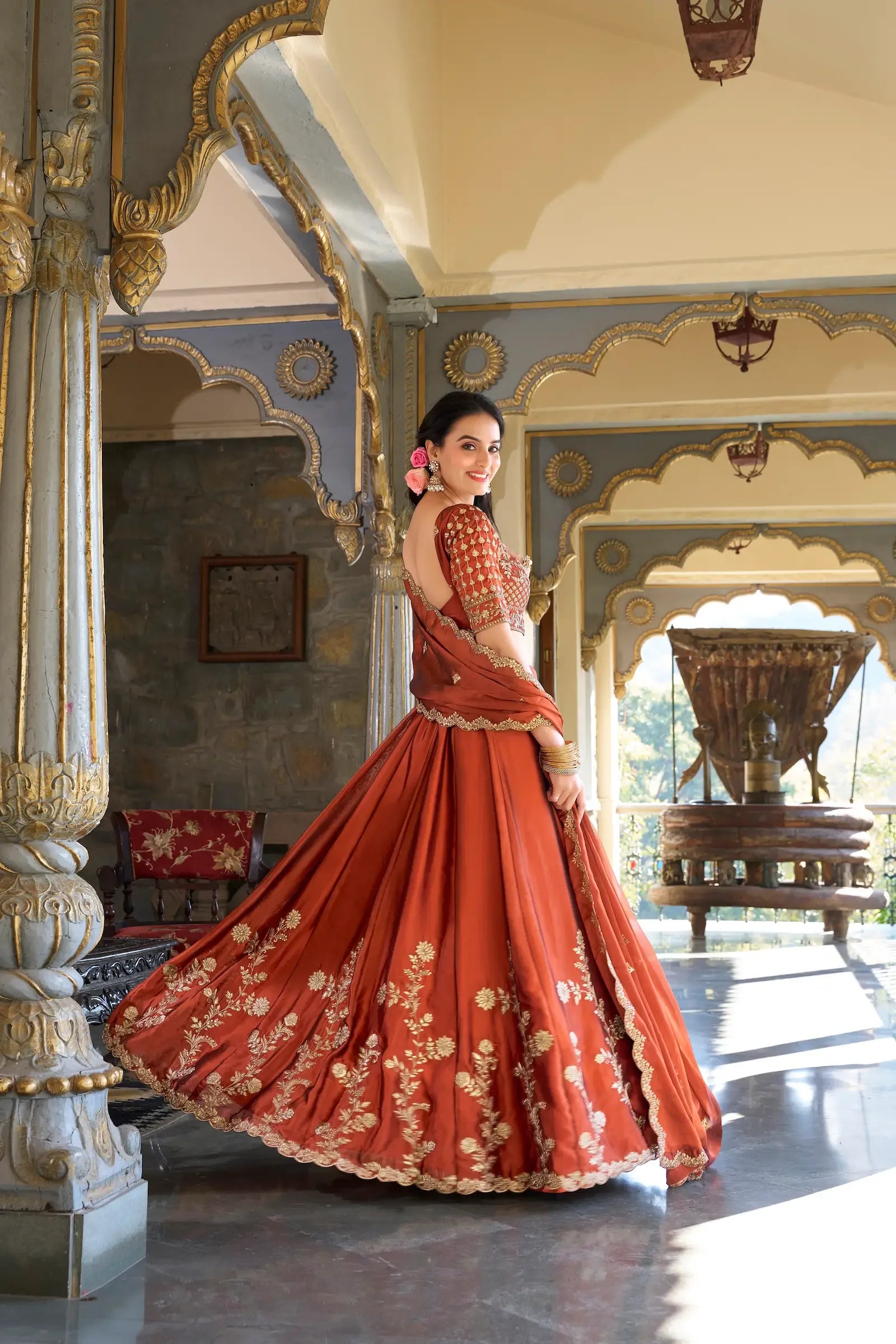 Woman in traditional rust orange embroidered lehenga twirling in ornate architectural hallway
